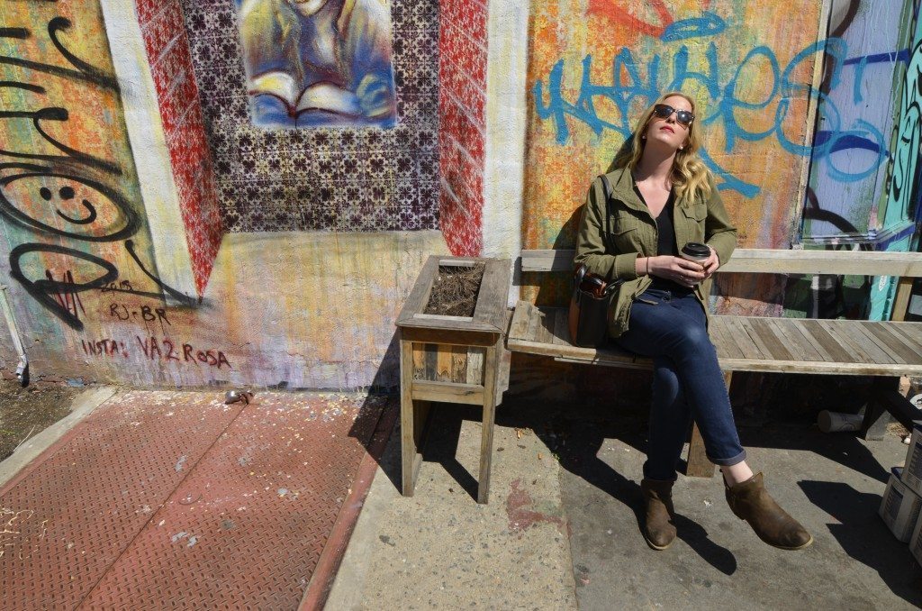 blonde woman with sunglasses and coffee in front of graffiti in the sunshine